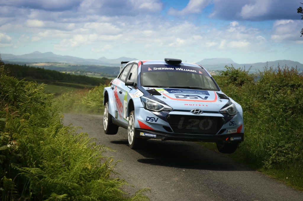 A rally car lifts off the road while speeding down a narrow country lane, surrounded by green hedgerows.