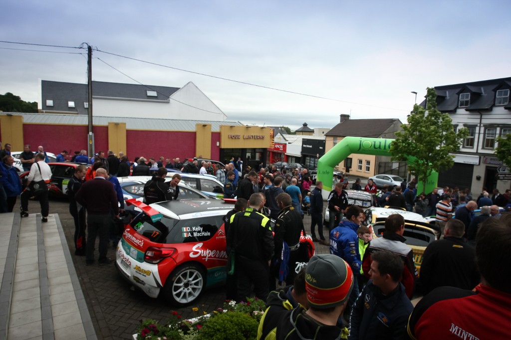 A crowd gathers in a town centre around rally cars at the starting line of the Donegal rally.