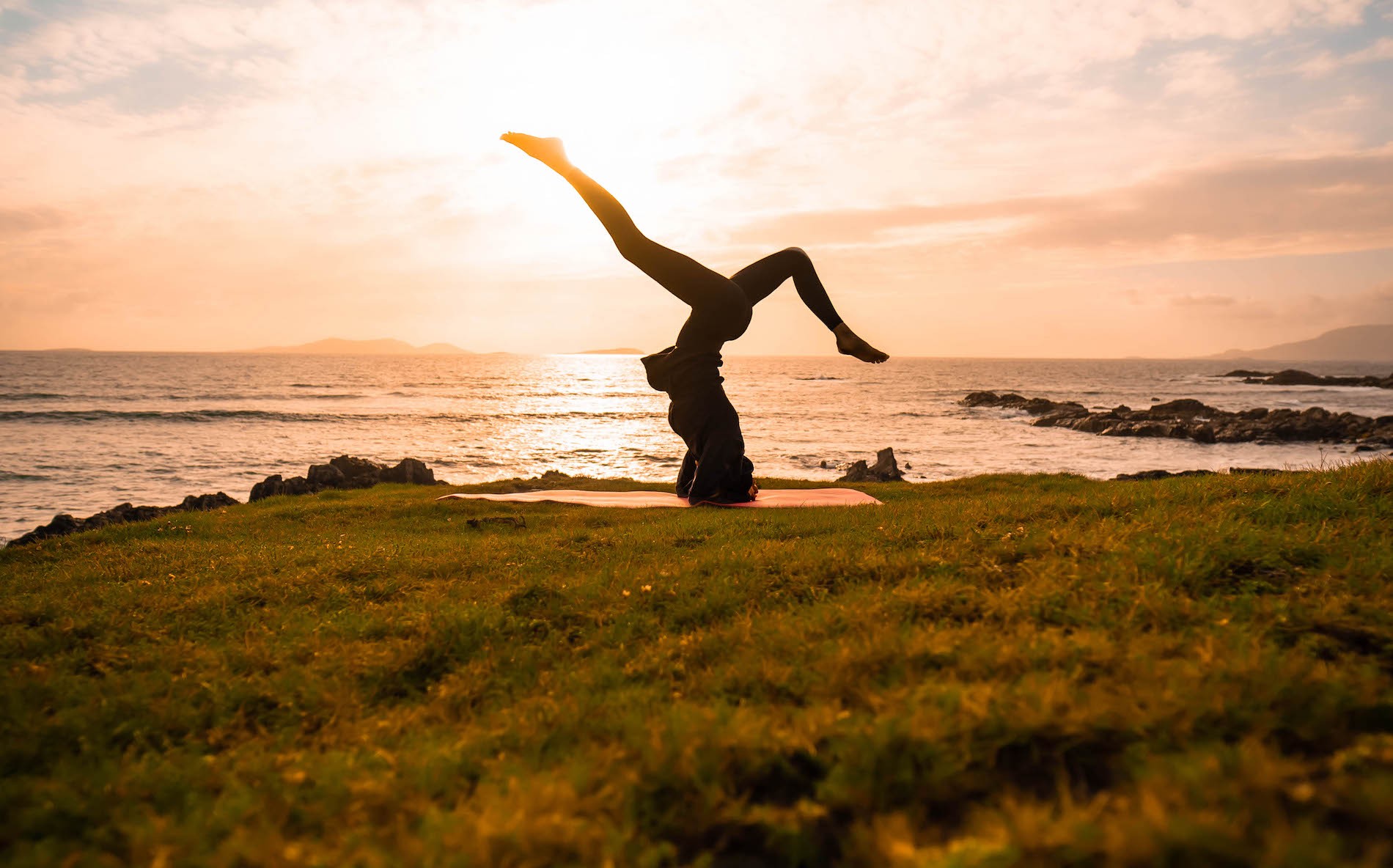 Person practicing yoga headstand on coastal grass at sunset with waves and rocks in background.