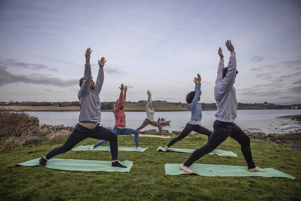 Group in warrior yoga pose on grass beside Strangford Lough under evening sky near anchored boat.