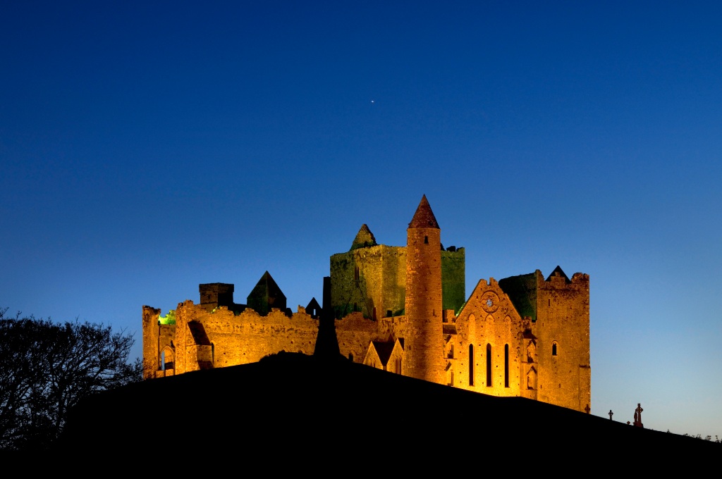 Rock of Cashel illuminated against a deep blue evening sky, with silhouettes in the foreground.