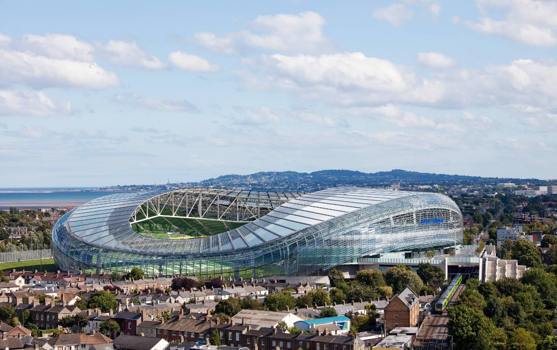 An aerial view of the modern glass-roofed Aviva Stadium in Dublin, nestled among city rooftops with the sea and hills in the distance.