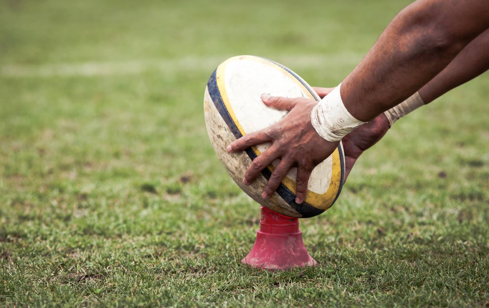 A close-up of a rugby player's hand placing a ball on a kicking tee during a match, set on a grassy field.