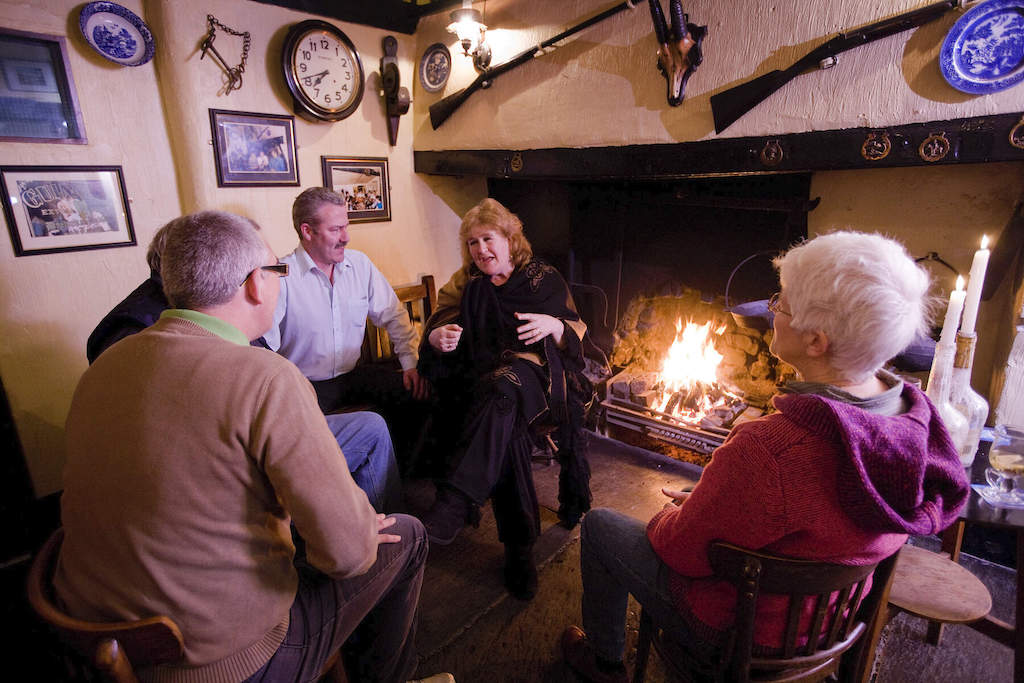 Group of friends chatting around a pub fireplace decorated with clocks and antiques in the Crosskeys Inn, County Antrim.