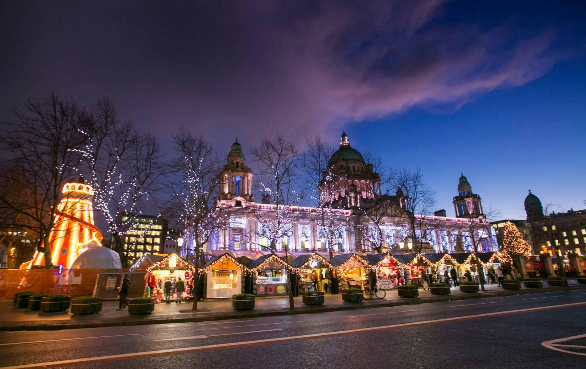 Belfast City Hall sparkles with festive lights above a lively Christmas market at dusk.