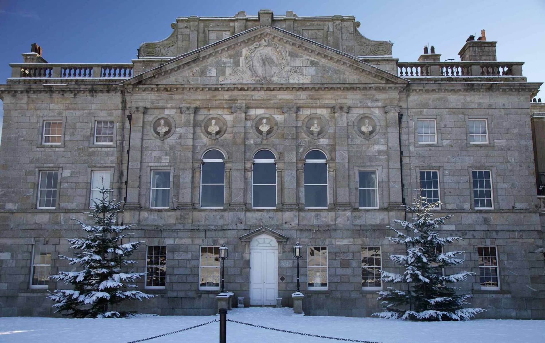 Snow-covered evergreens flank the grand facade of Powerscourt Estate in a crisp winter scene.