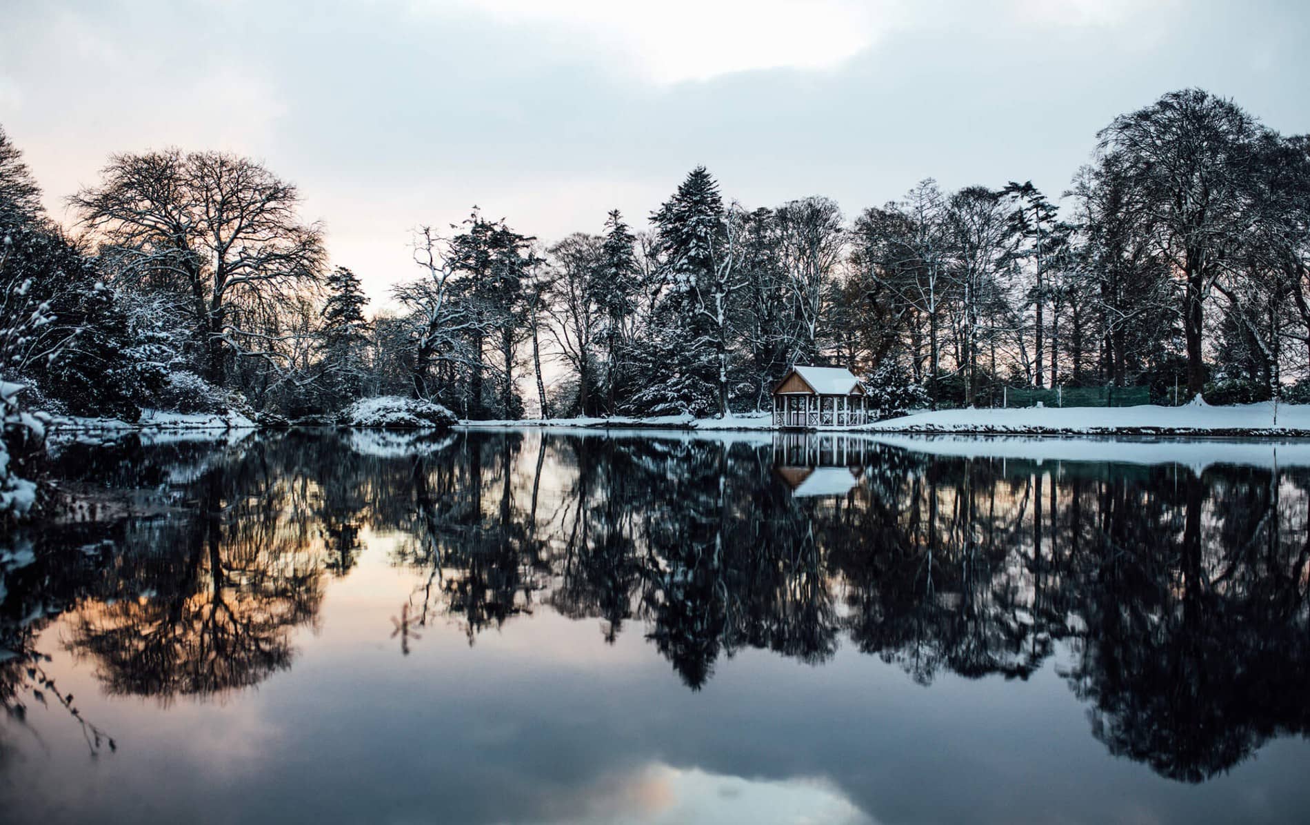A snowy lakeside hut is mirrored in still waters beneath frost-draped trees at dusk.