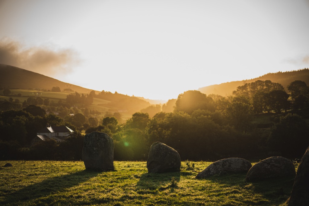 Low stone circle on grassy land at sunset in County Wicklow, with silhouettes of trees and hills in the background.