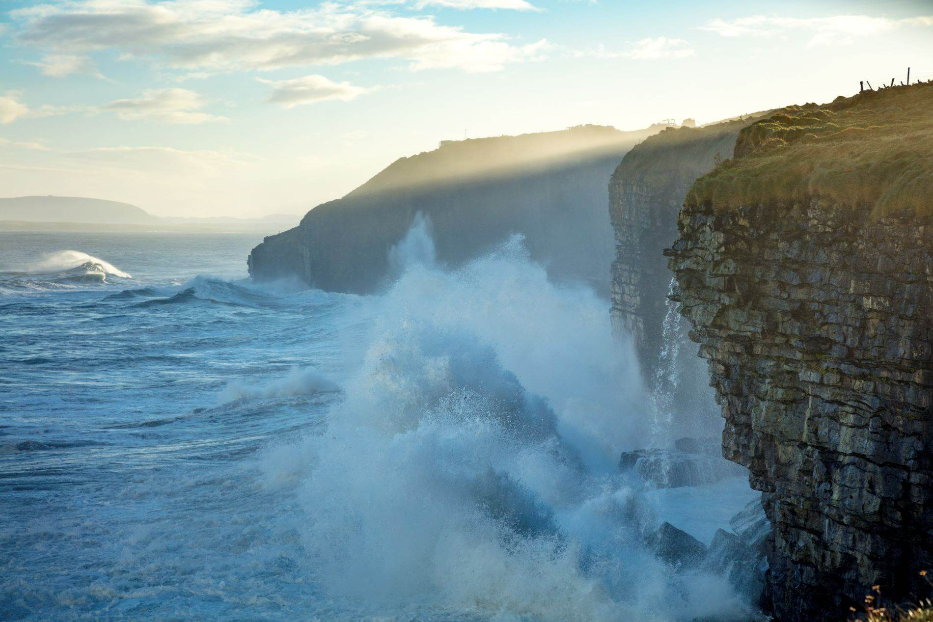 Dramatic Atlantic waves crash against high sea cliffs in County Sligo at golden hour, with sunlight breaking through mist.