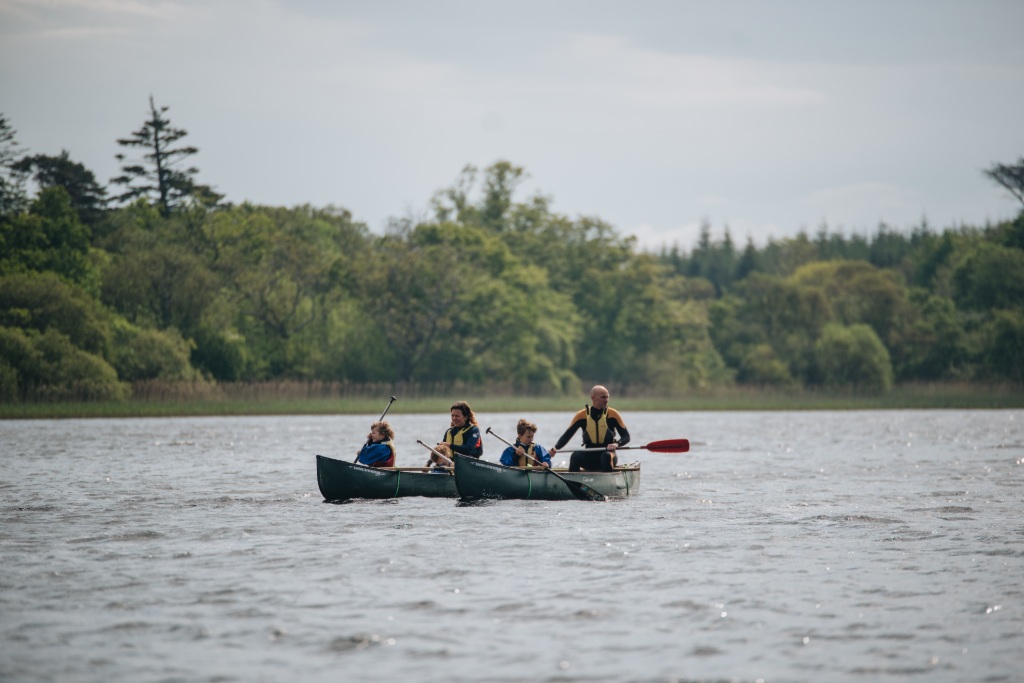Two canoes with a family of five paddle across a lake in County Sligo, with dense forest lining the lakeshore.