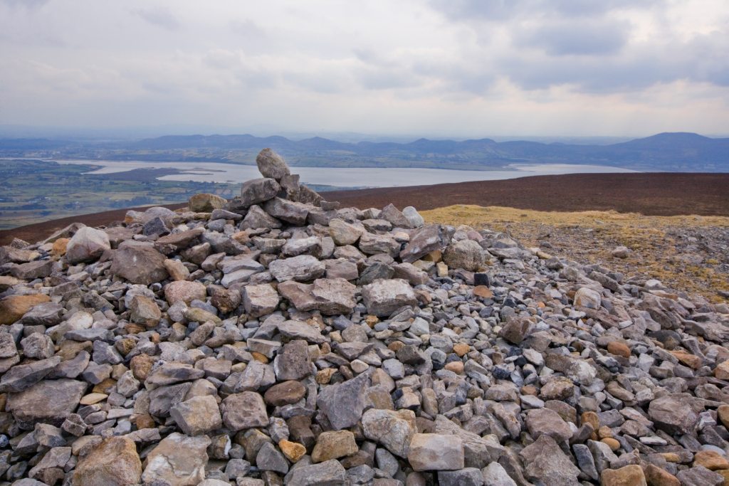Stone cairn atop Knocknarea mountain overlooks coastal inlets and countryside in County Sligo under overcast skies.