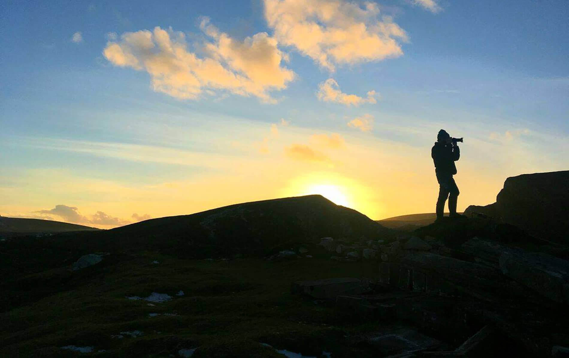 Silhouette of a person with a camera standing on a rocky hilltop in Portacloy, County Mayo, during a golden sunset.