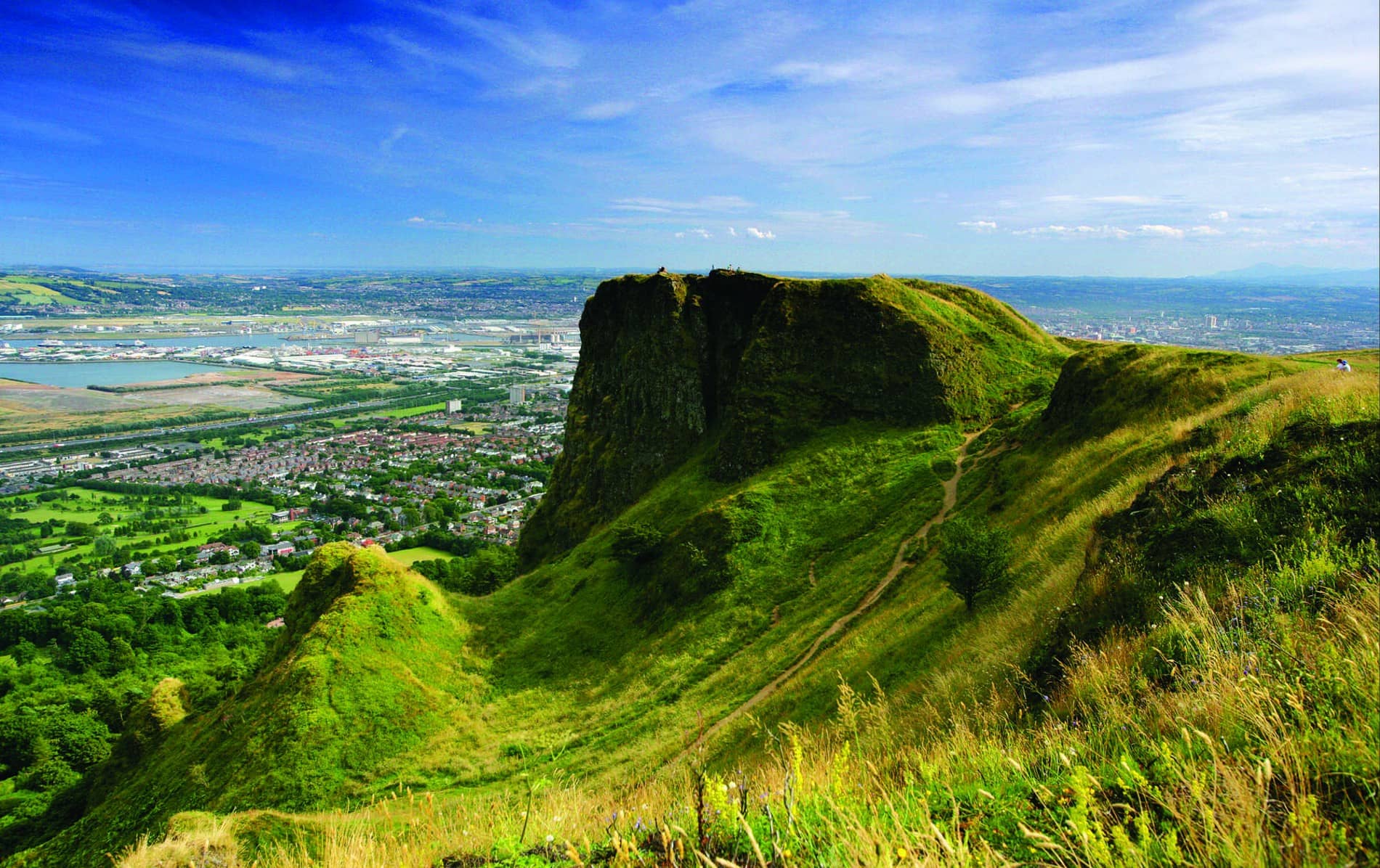 Green slopes of Cave Hill overlook Belfast city, with panoramic views of urban sprawl, coastline and distant hills on a sunny day.