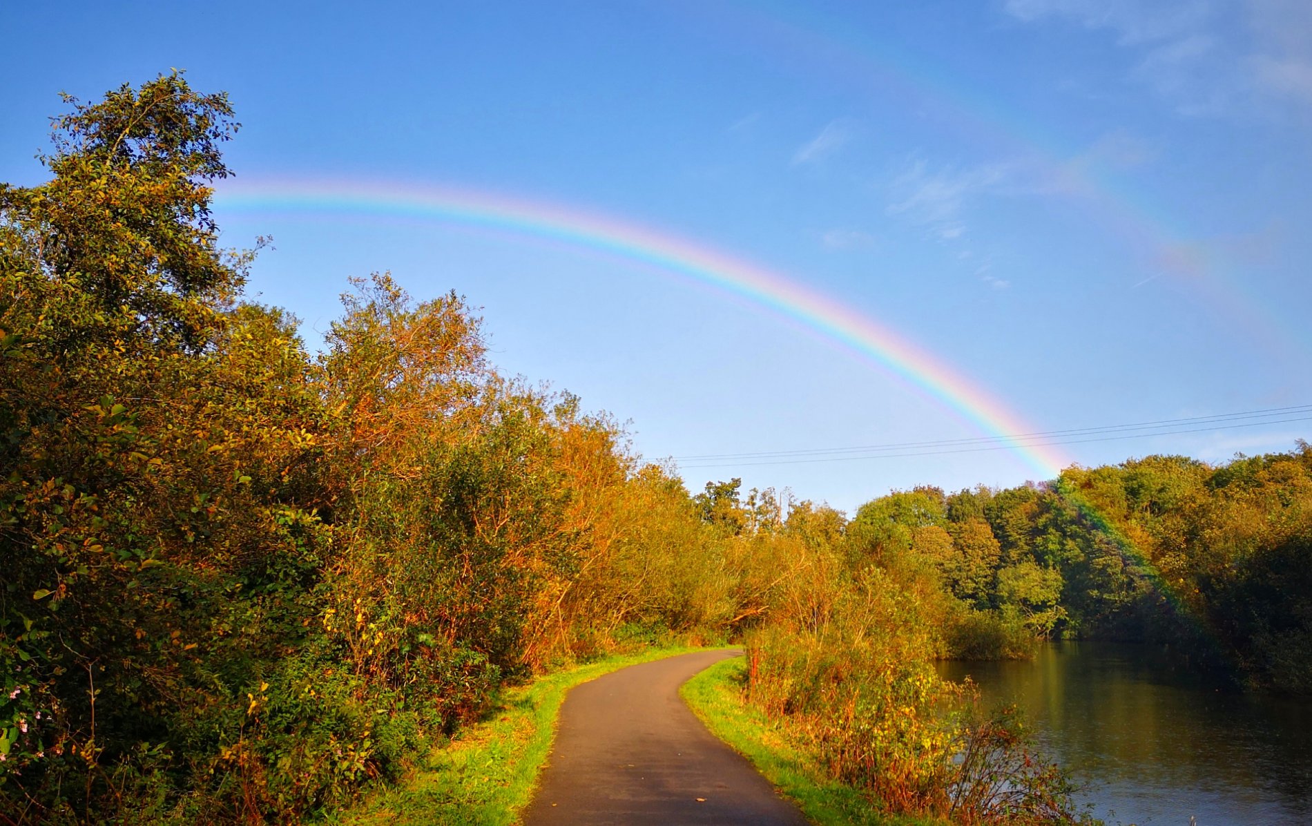 A rainbow arcs above a quiet towpath beside the River Lagan in Belfast, lined with autumn trees under a clear blue sky.