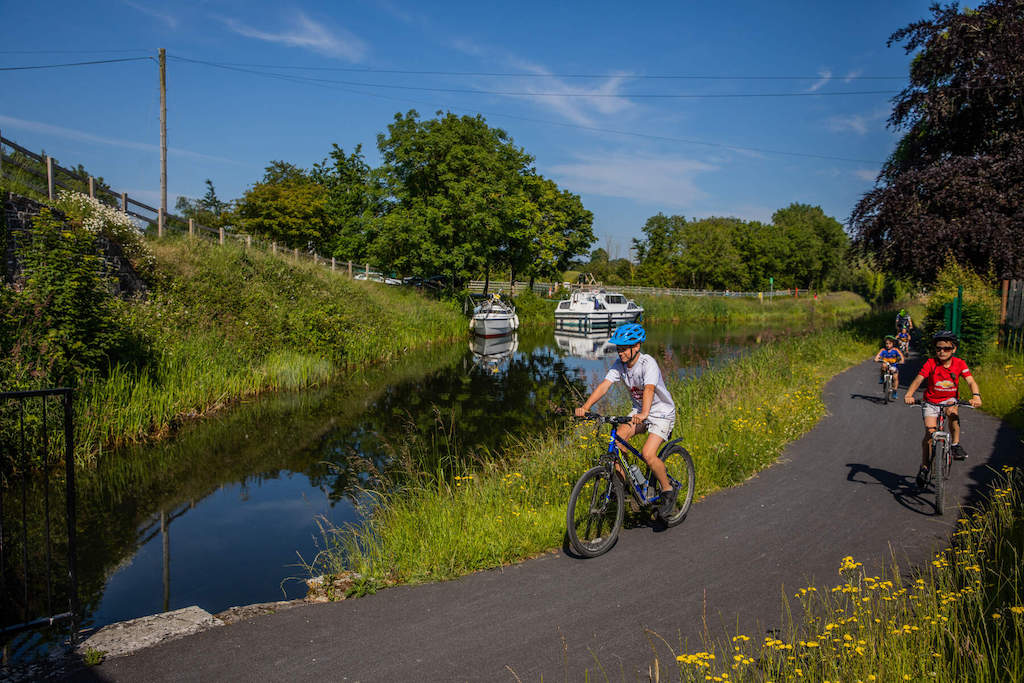 Children cycling along the Royal Canal Greenway on a sunny day, with boats moored on the water nearby.