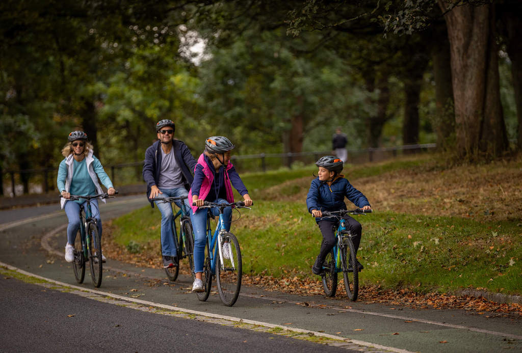 Family of four cycling together on a leafy path in the Phoenix Park, Dublin.