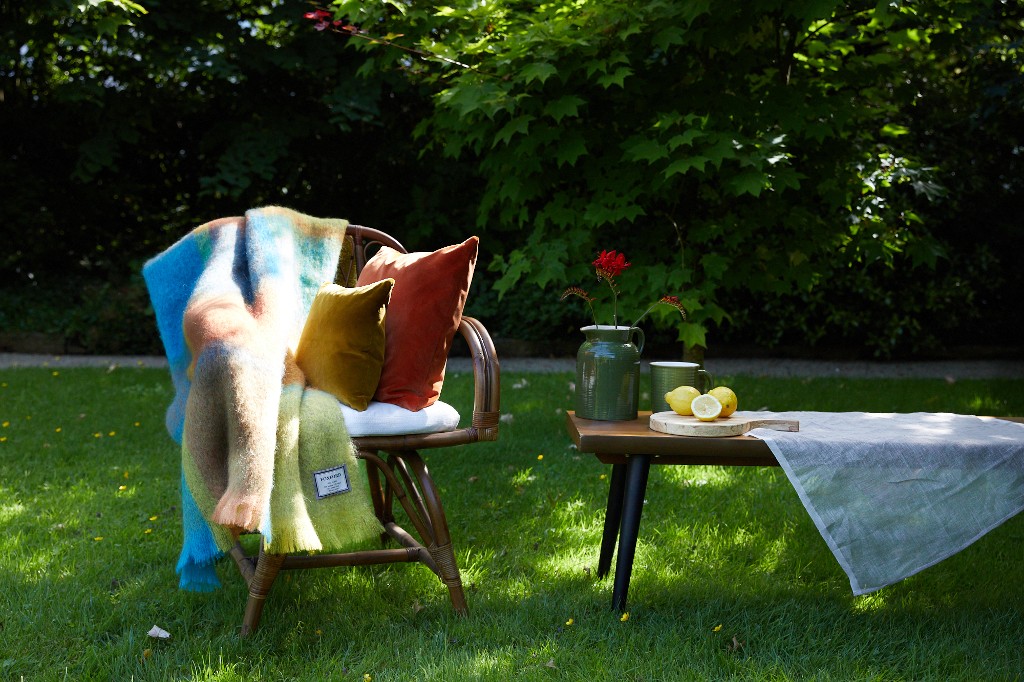 Colourful mohair throw draped over a wicker chair with cushions beside a garden table set with lemons and a vase.