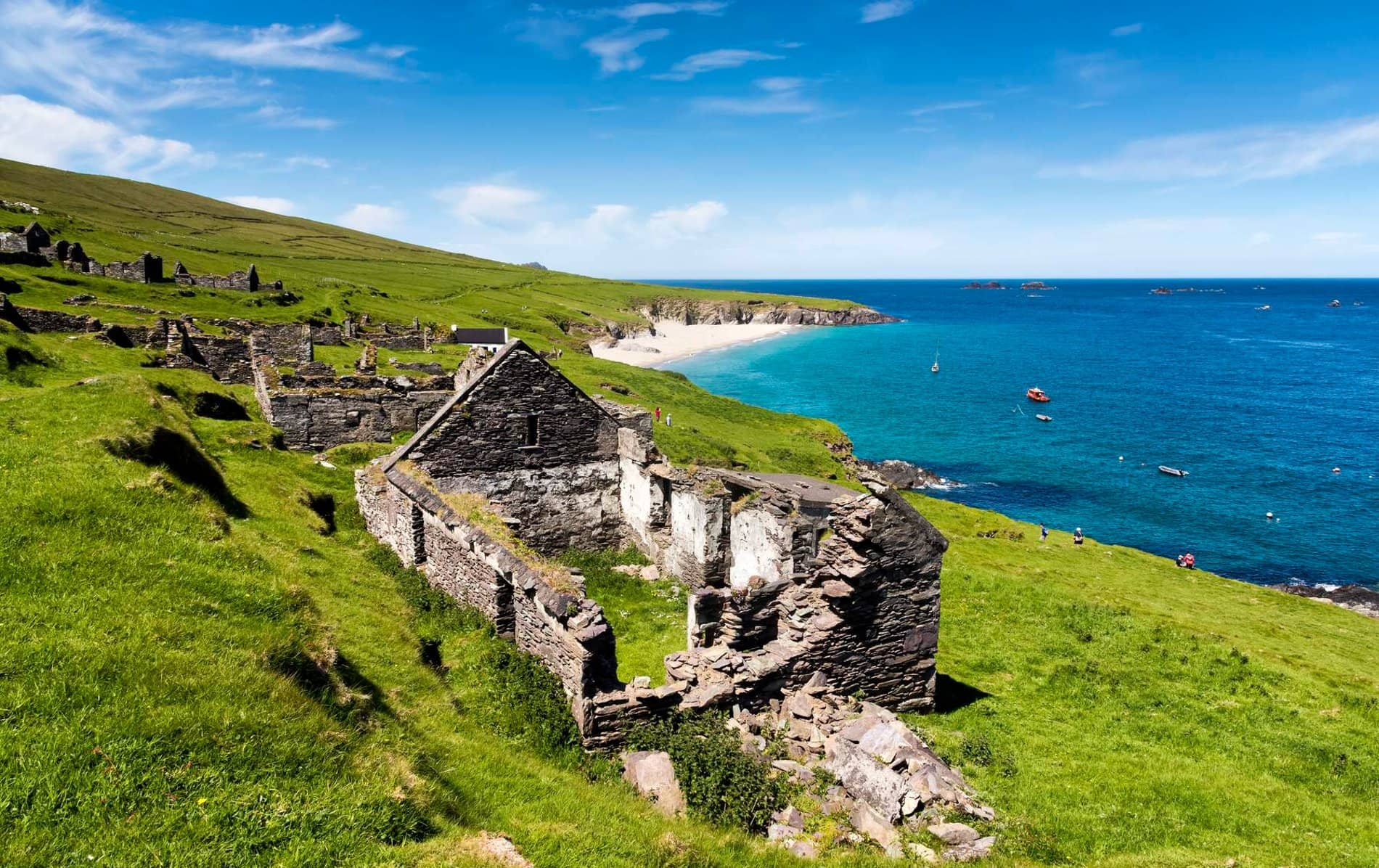 Stone ruins of a house on Great Blasket Island overlook grassy cliffs and a clear blue sea dotted with small fishing boats.