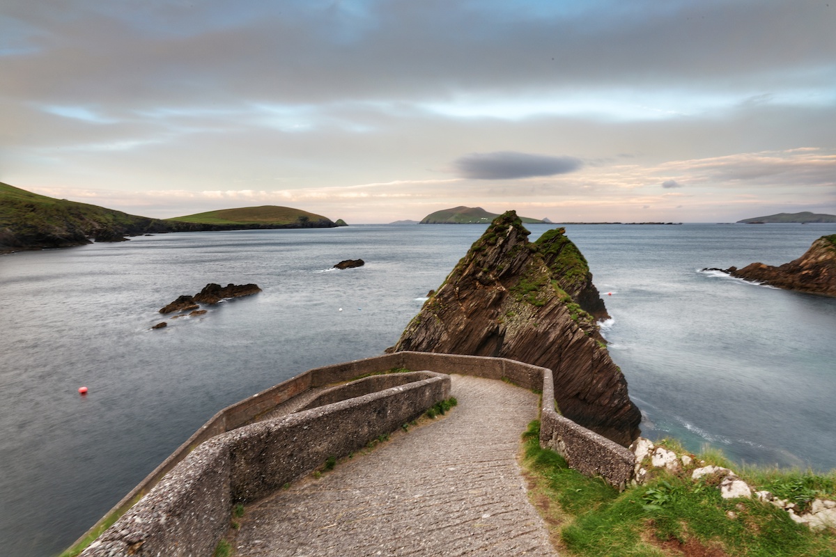 Curved stone pier at Dunquin Harbour leading to the sea, with sharp rocky outcrops and the Blasket Islands in the distance.