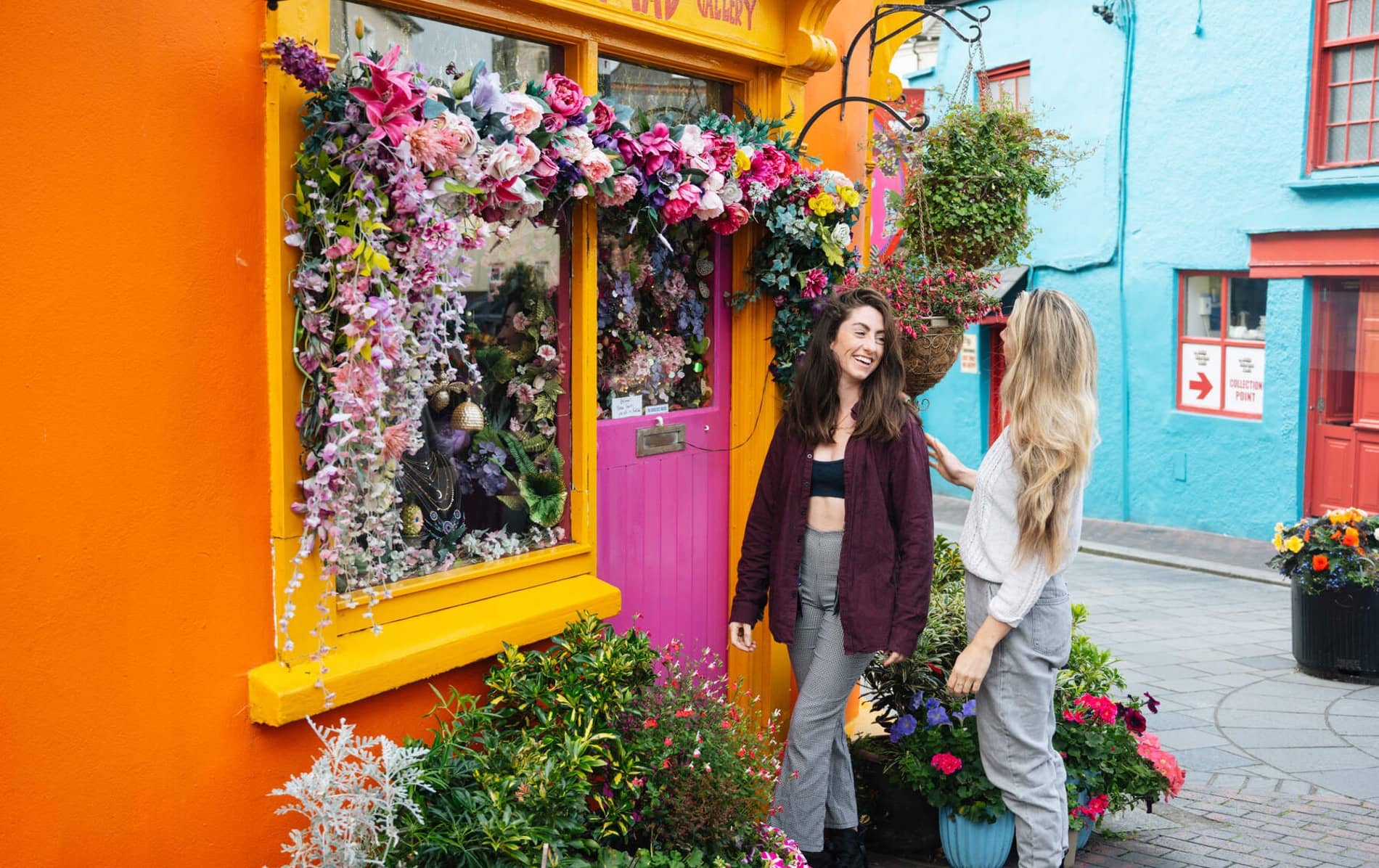 Two women chatting outside a flower-decorated shopfront on a colourful street in Kinsale, County Cork.