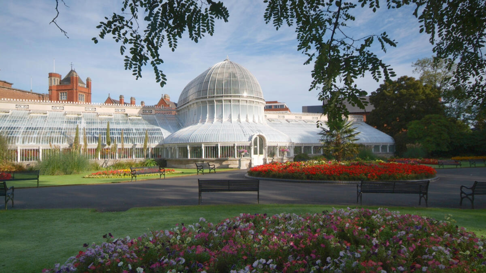 Victorian domed glasshouse in Belfast Botanic Gardens, framed by flowerbeds and benches on a bright day.