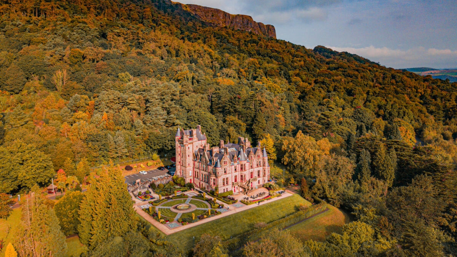 Belfast Castle nestled in autumn woodland on the slopes of Cave Hill, glowing in warm sunset light above the Belfast landscape.