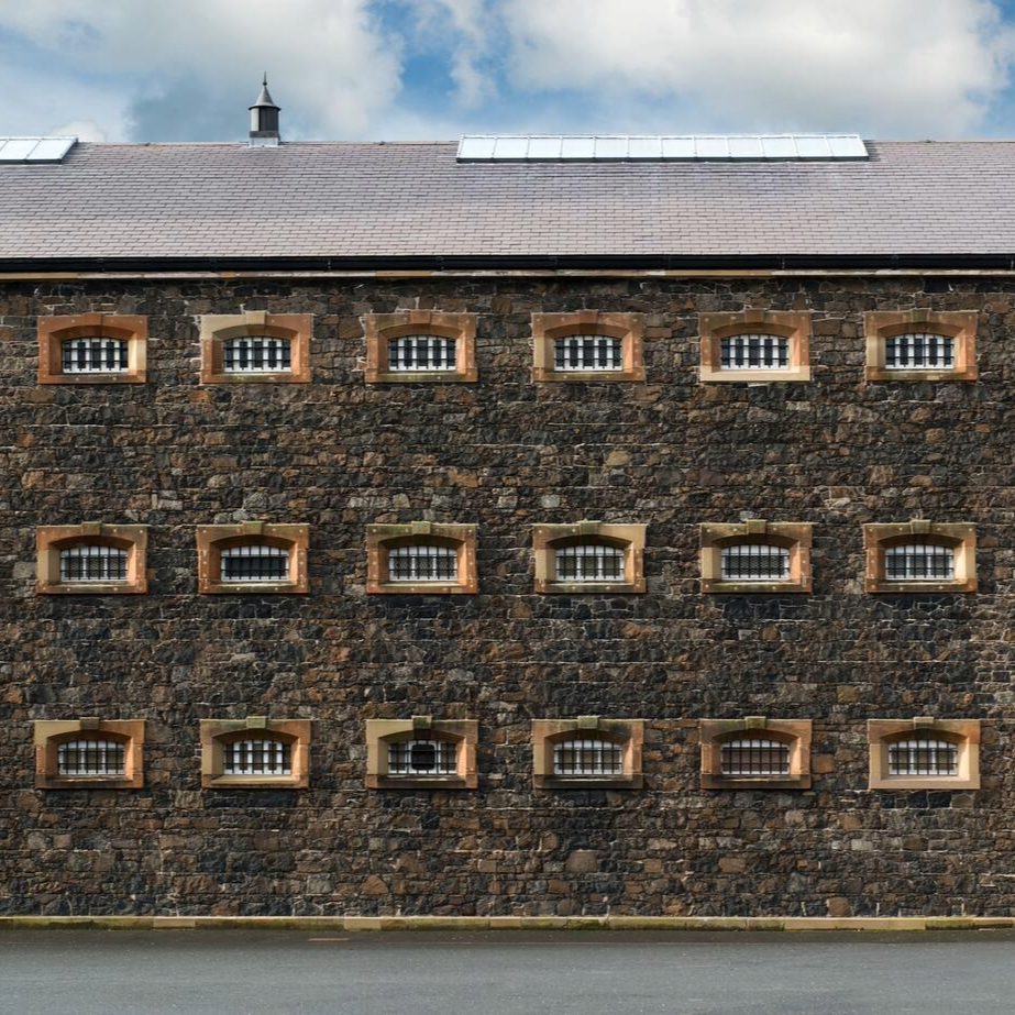 Stone exterior of Crumlin Road Gaol in Belfast, with rows of barred windows set into a dark stone façade.