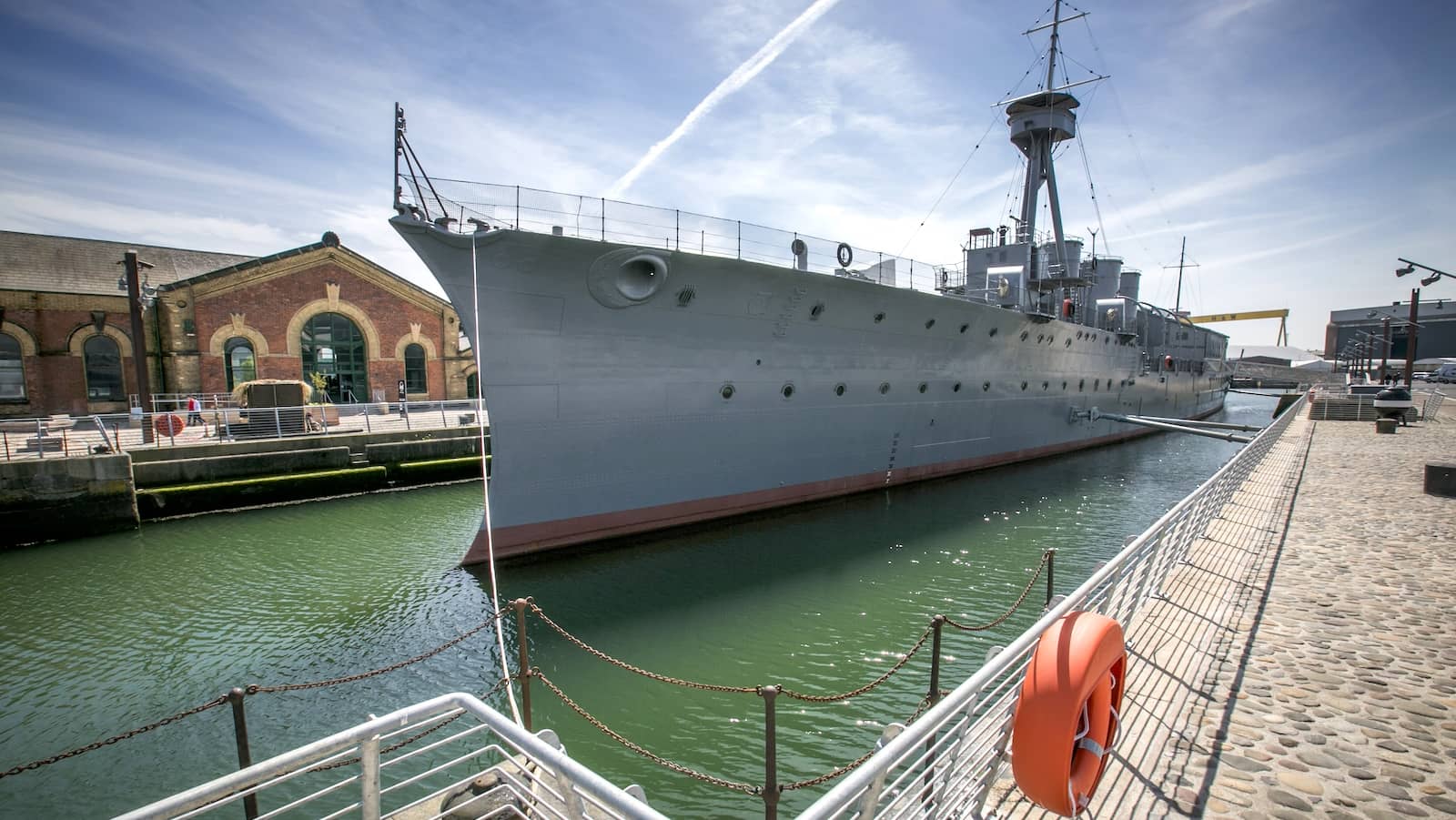 HMS Caroline moored at Titanic Quarter, Belfast, with historic brick buildings and harbour walkway.