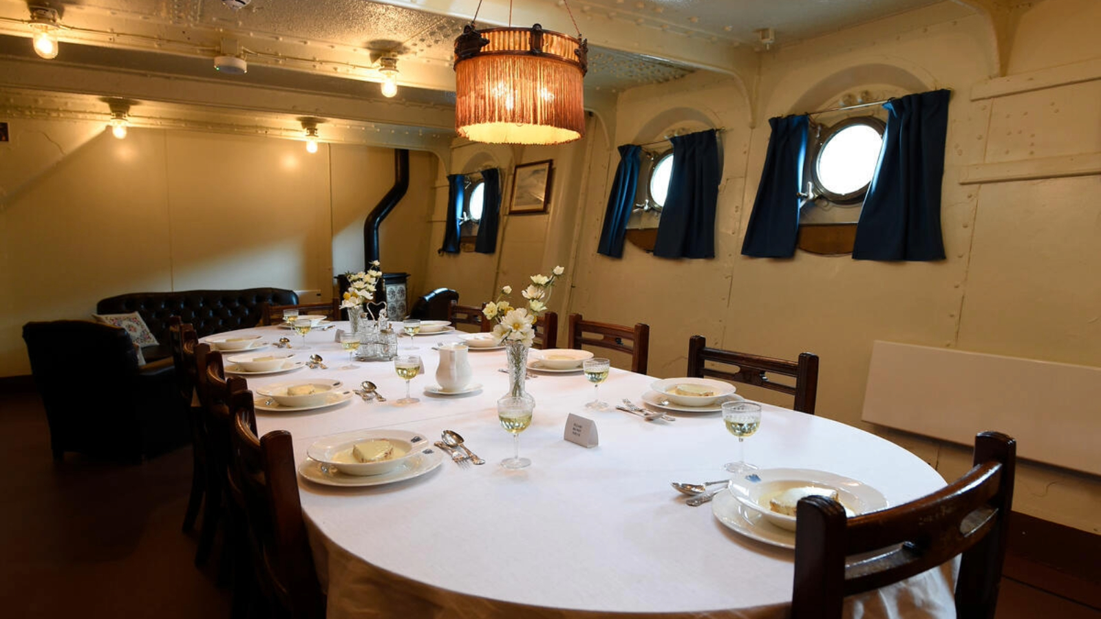 Dining room set for a meal on HMS Caroline with white tablecloths, place settings and porthole windows.