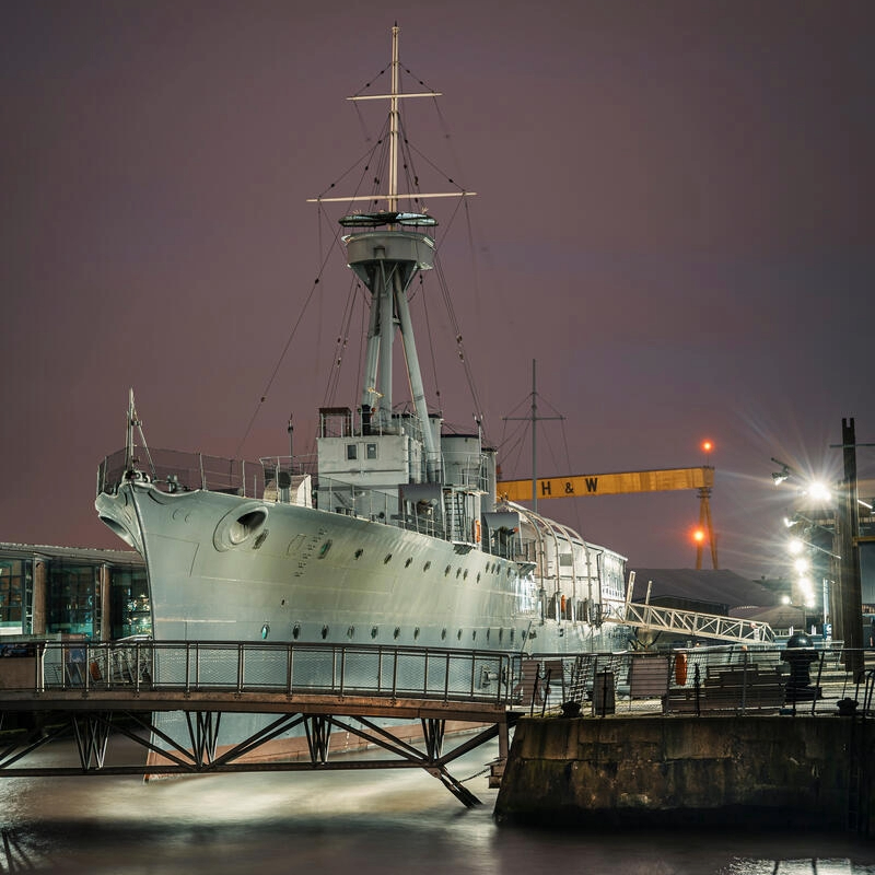 HMS Caroline moored in Belfast's Titanic Quarter at night with Harland & Wolff cranes in the background.