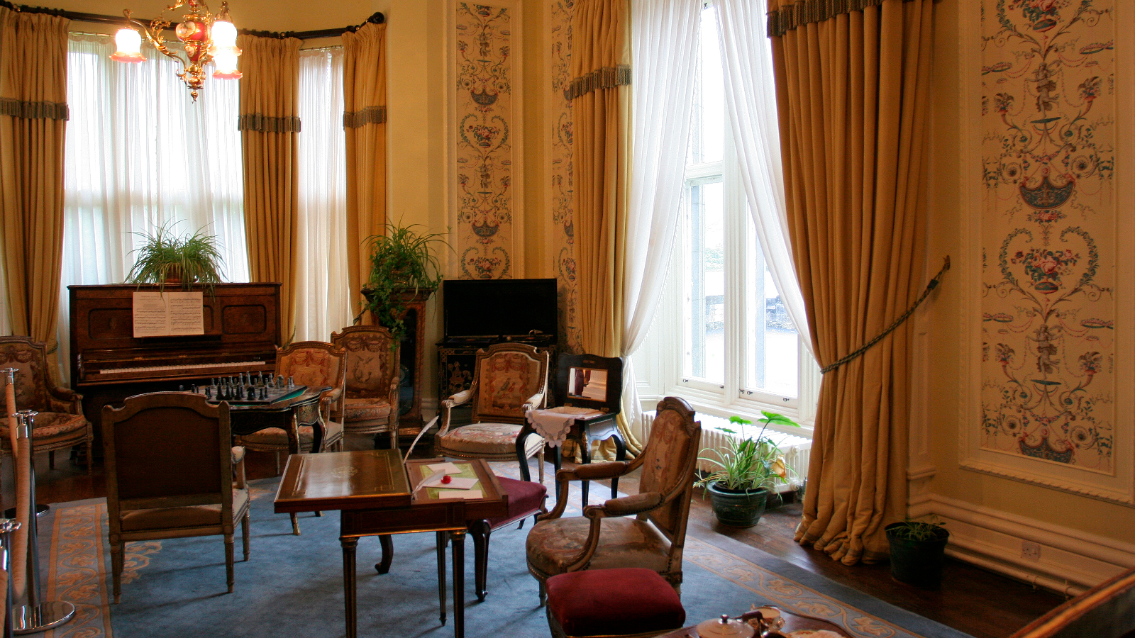 Elegant Victorian room inside Kylemore Abbey with piano, chess table and tall draped windows.