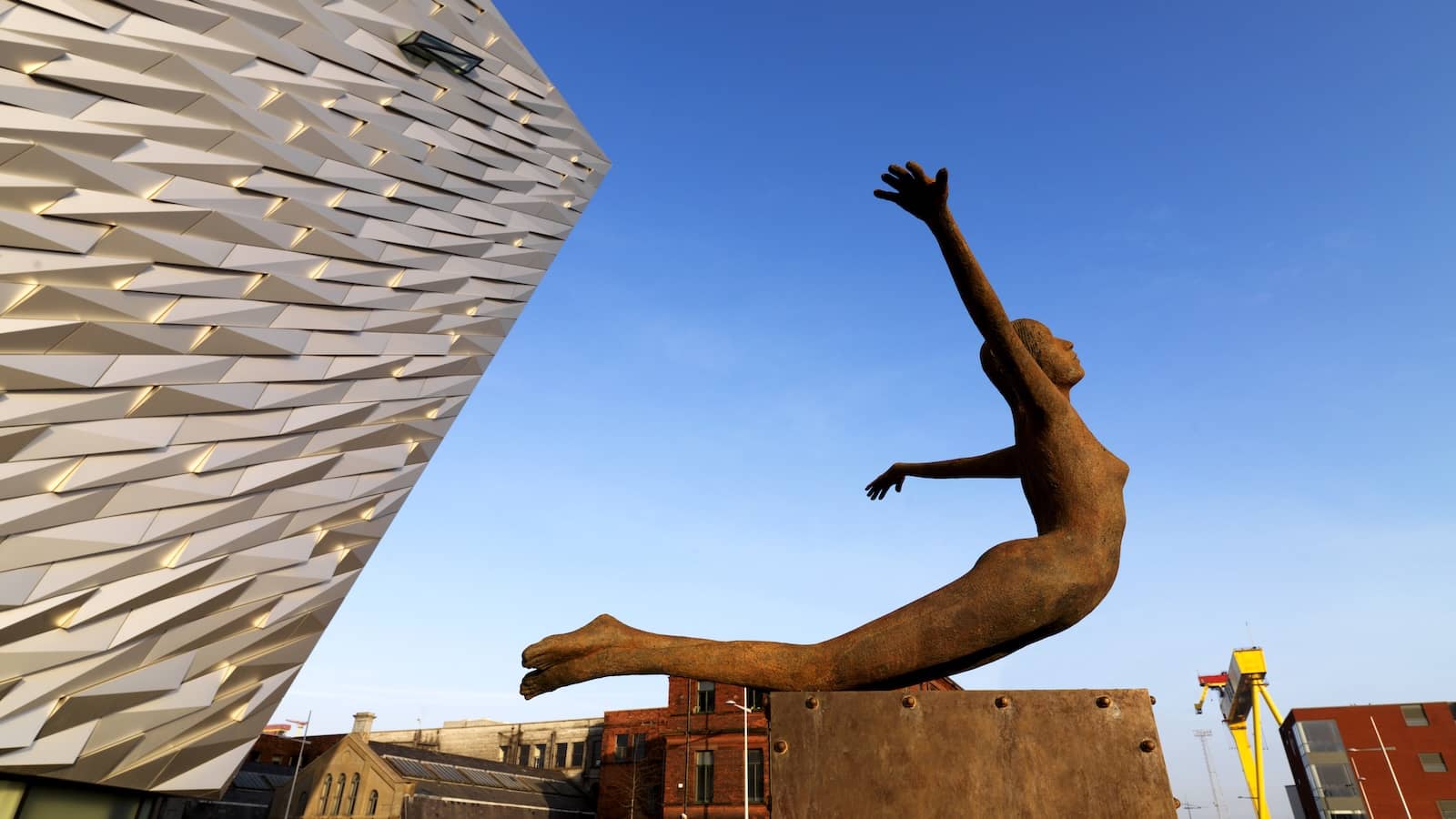 Rust sculpture of a diving figure beside the angular façade of Titanic Belfast under a clear blue sky.