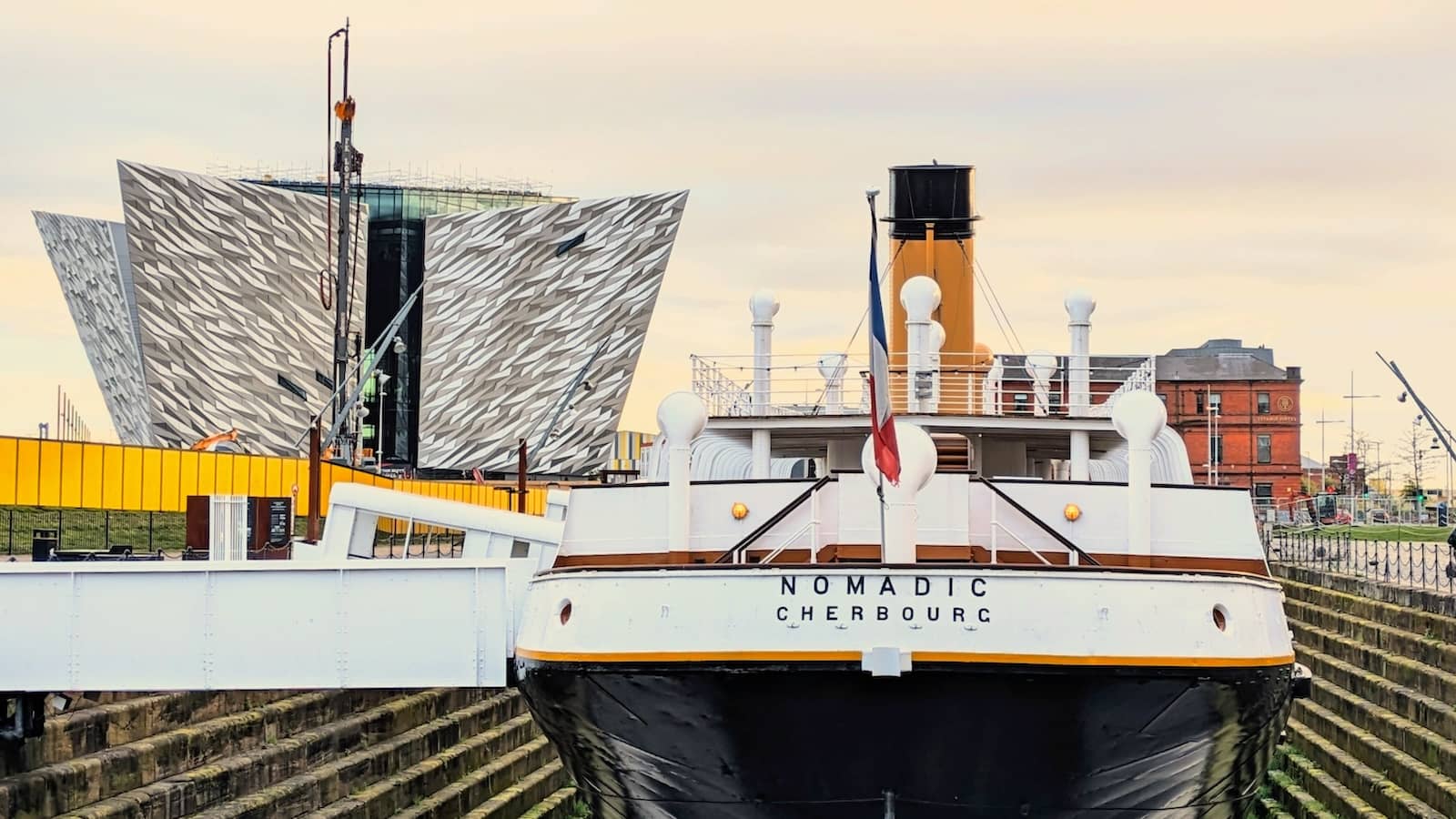 SS Nomadic moored in dry dock with the modern Titanic Belfast building rising behind it.