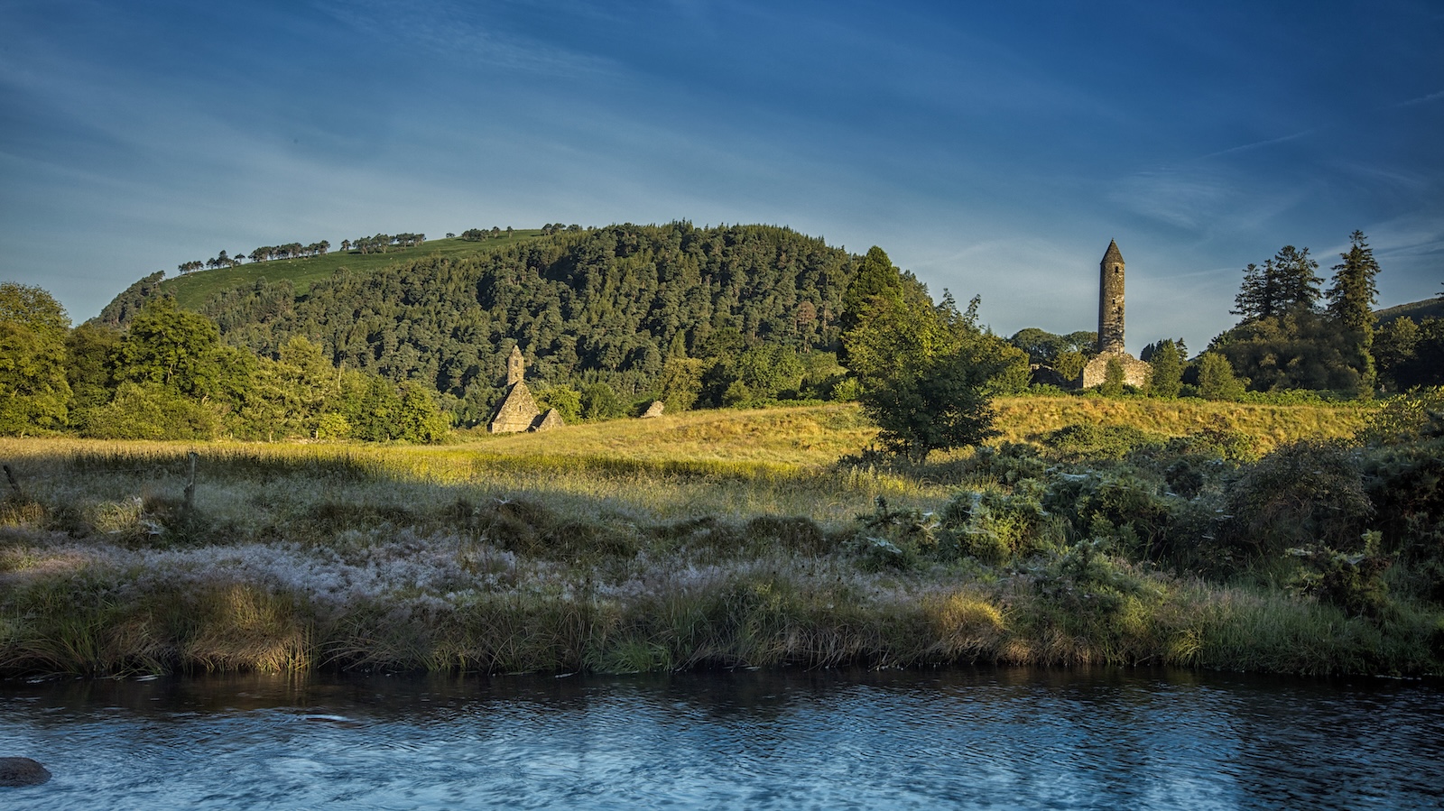 Round tower and stone church beside meadow and river at Glendalough in Wicklow Mountains National Park.