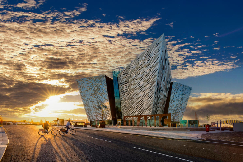 Two cyclists heading towards Titanic Belfast as the sun rises against a blue sky with some clouds.
