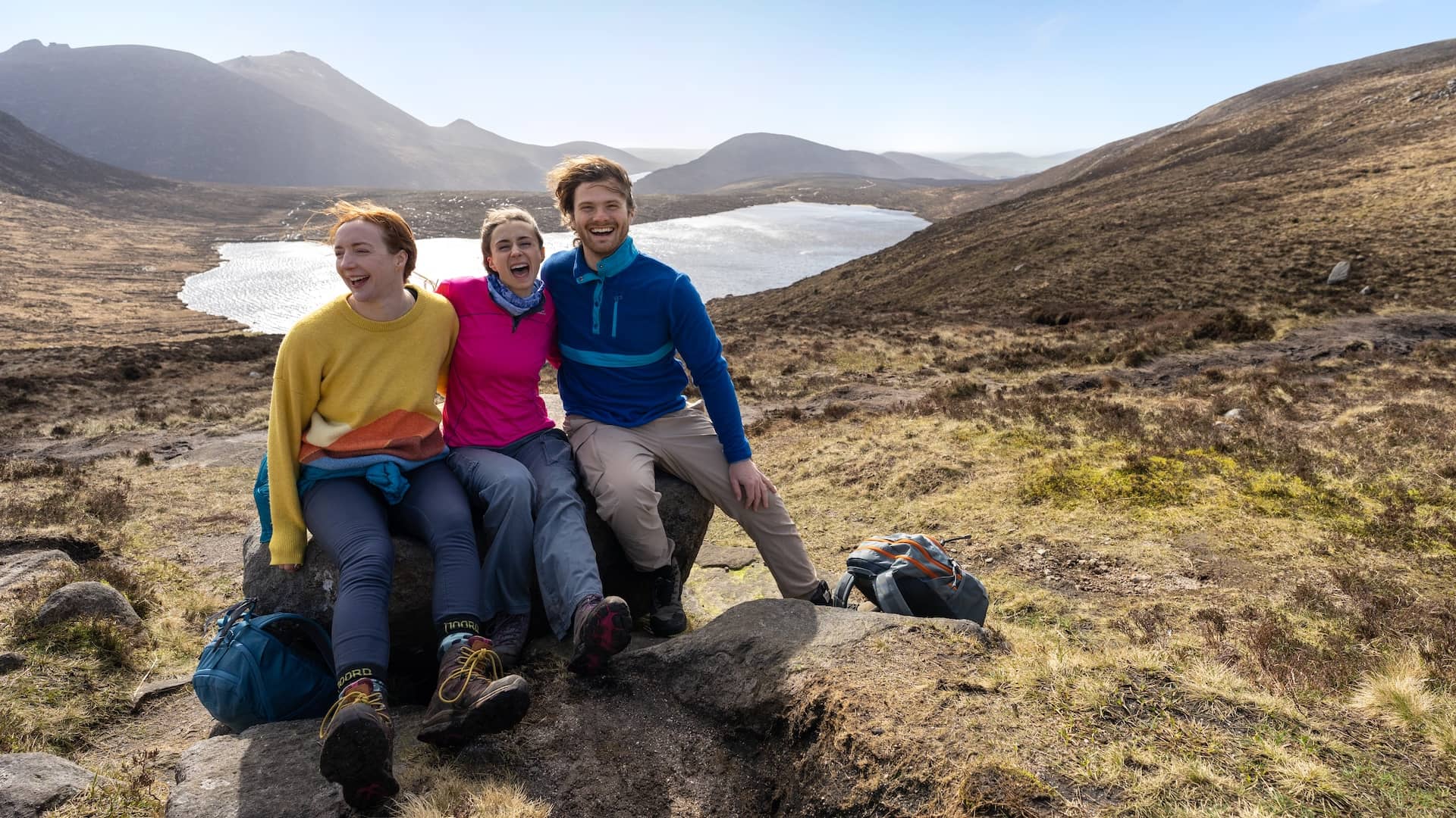 Three friends resting on a rocky outcrop in the Mourne Mountains, County Down, with sweeping views over a lake and rugged peaks.