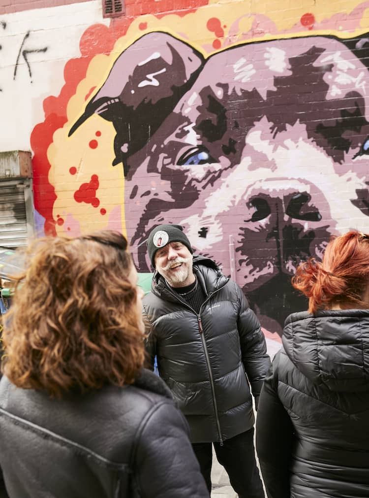 Guide chatting with visitors beside a vibrant dog mural in a Belfast alleyway during a street art tour.