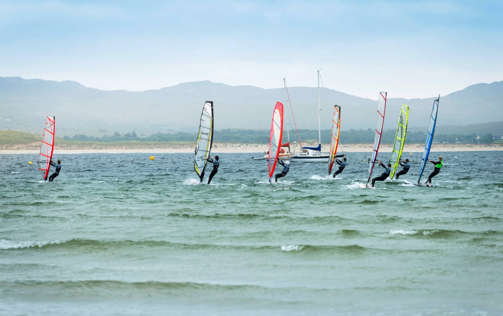Windsurfers glide across choppy waves near the coast in County Donegal, with sails lit by soft summer light.