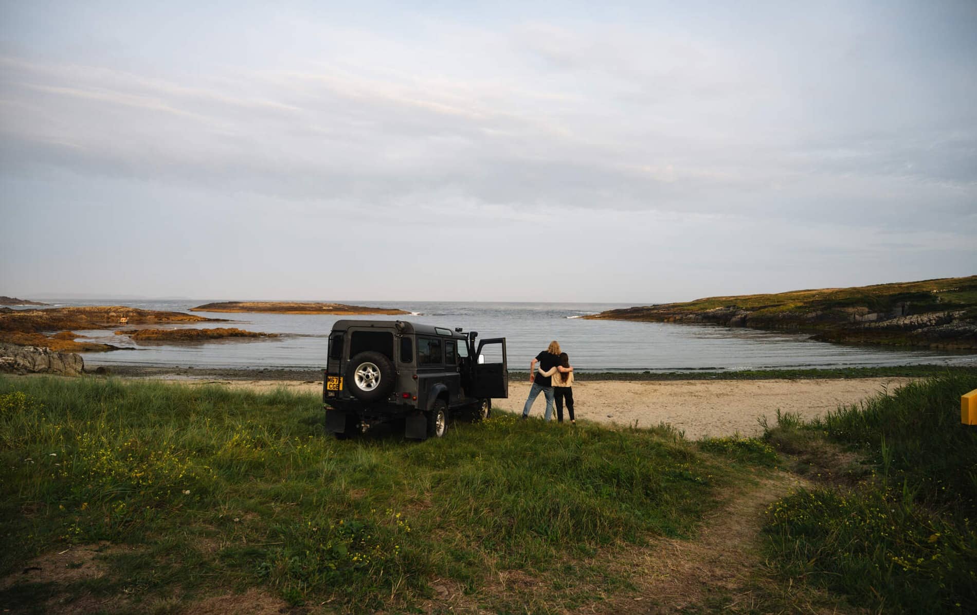 A couple embraces beside a black jeep parked on a quiet beach cove at Crookhaven, Cork.