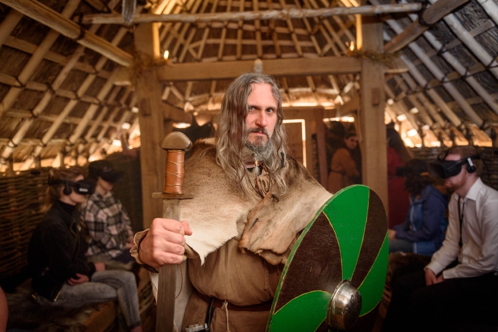 Viking reenactor holding sword and green shield inside a reconstructed hall with visitors watching.