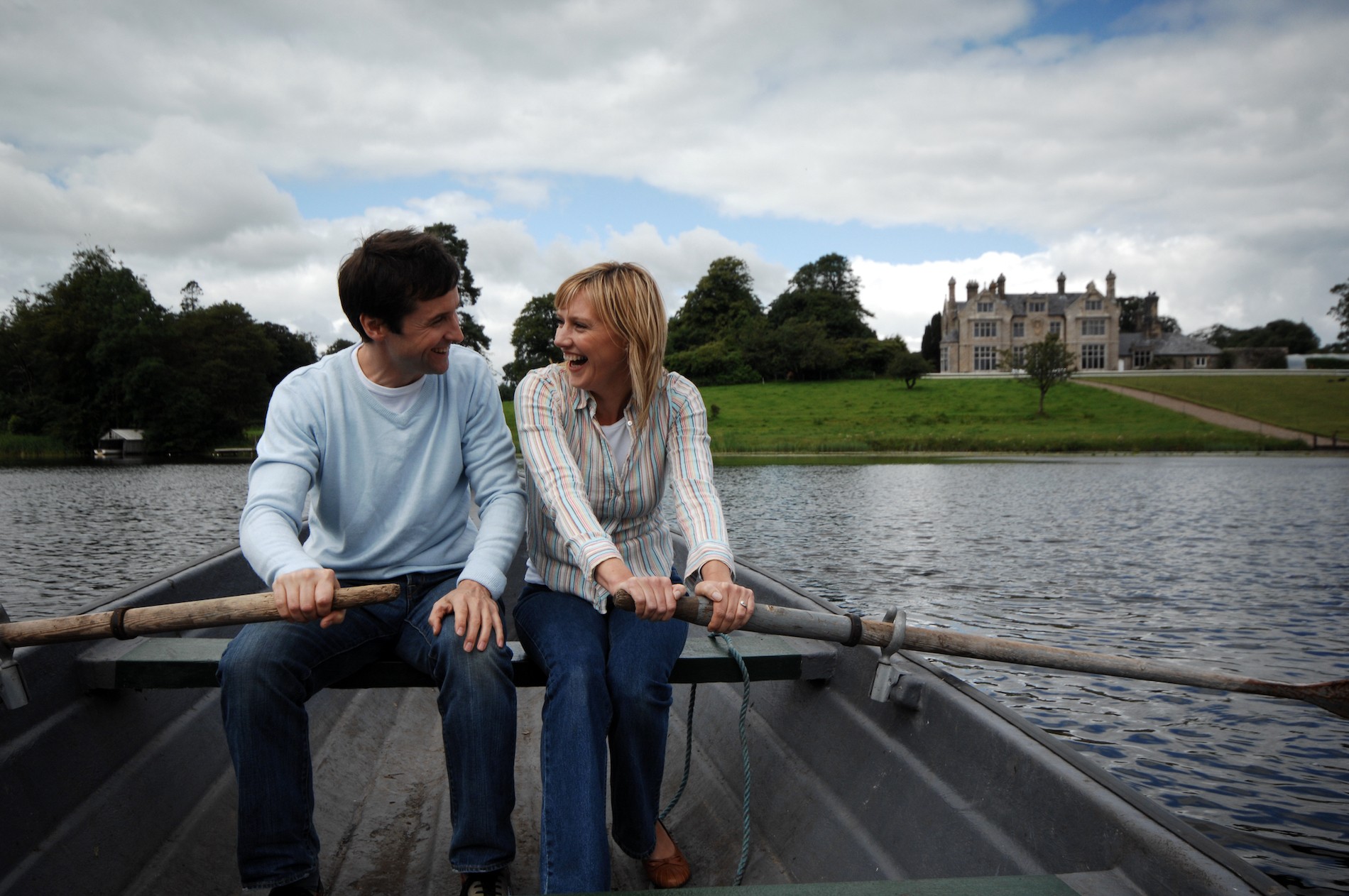 Couple laughing in a rowboat on a lake with a stately home and lush green grounds behind, promoting wedding adventures.