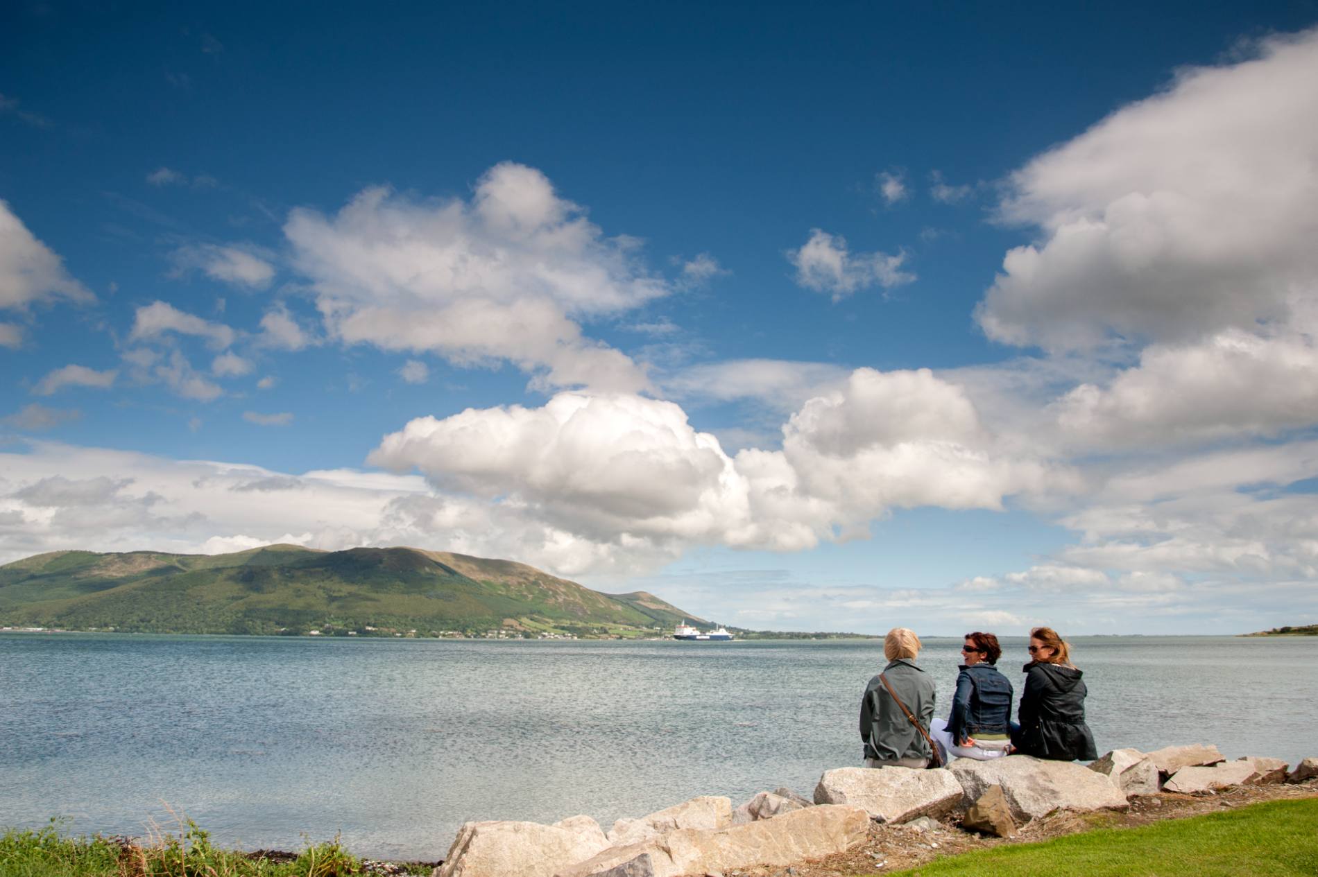 Three women walking on the coast with mountains and blue sky in the background.