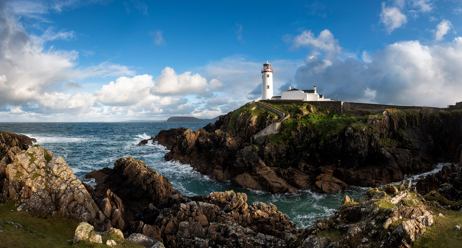 White lighthouse at Fanad Head stands above crashing waves and rugged cliffs under a bright blue sky.