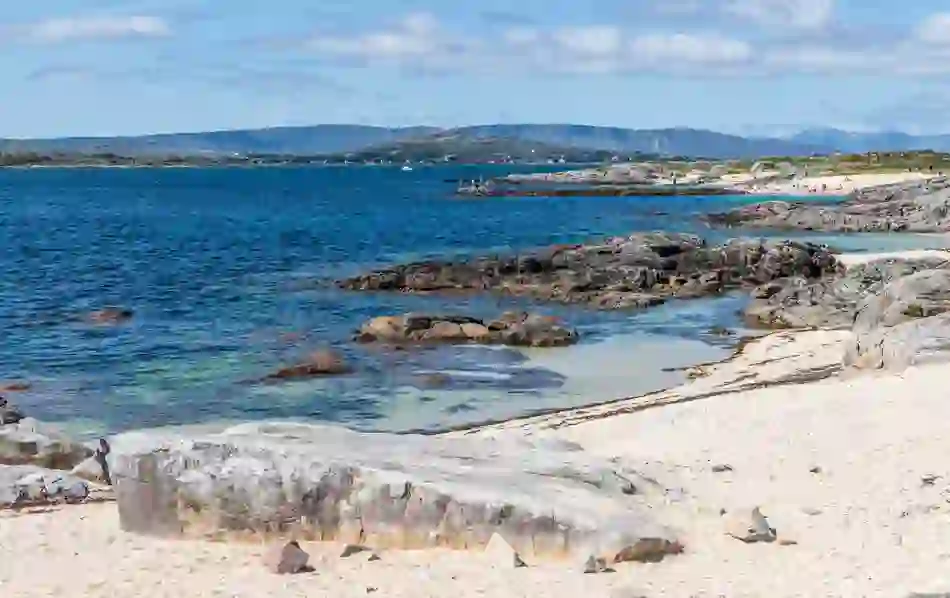 Clear blue waters and white sand shoreline at Carraroe Beach, Galway, with rocky outcrops and hills beyond.