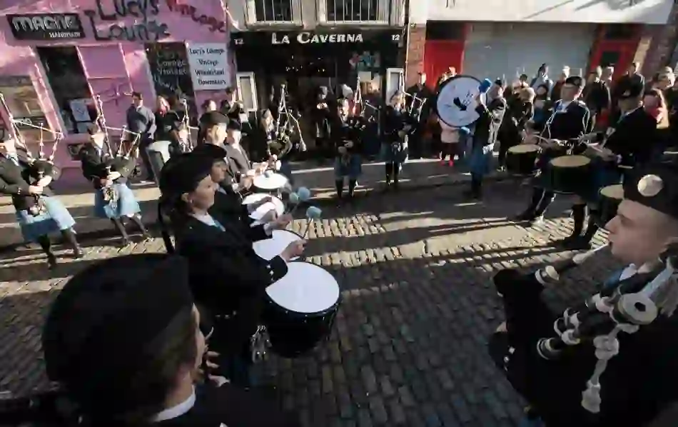 Pipe band musicians perform on a cobbled street in Temple Bar, Dublin, watched by a crowd in winter clothing.