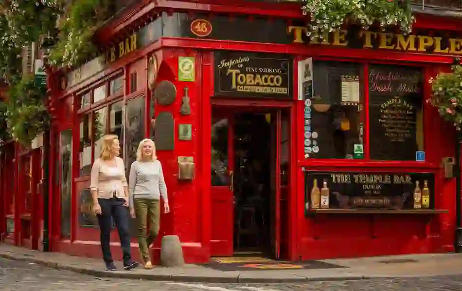 Two women walk past the iconic red façade of The Temple Bar pub in Dublin, known for traditional Irish music.