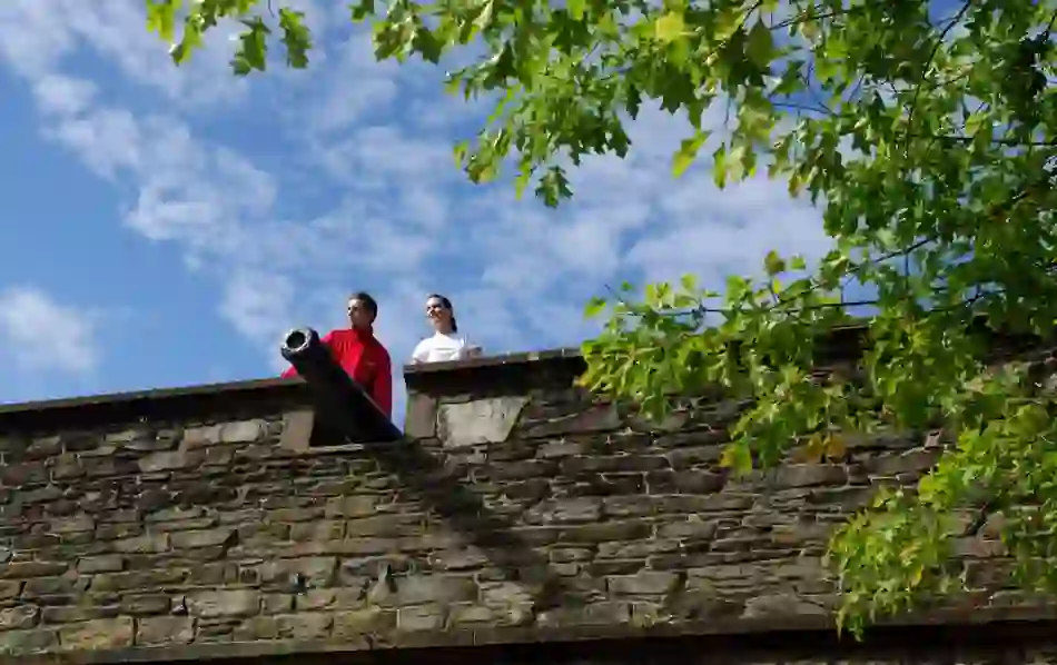 Couple stands by cannon on Derry~Londonderry's city walls beneath leafy trees and blue sky.