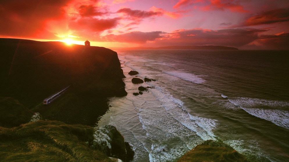 Sunset behind Mussenden Temple on a cliff in Northern Ireland, with waves crashing below and a red-tinted sky above.