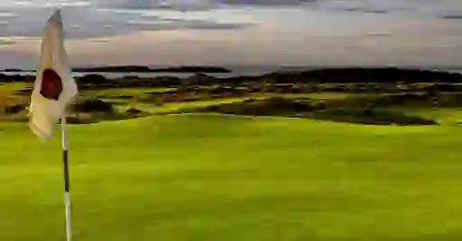 A golf flag stands on lush fairways overlooking the Atlantic Ocean, with golden evening light on rolling dunes.