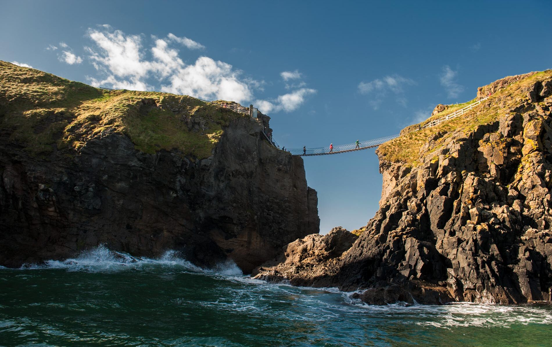 Visitors cross the Carrick-a-Rede Rope Bridge between sea cliffs in County Antrim on a sunny day.